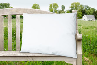 Eine weißes Kopfkissen ist auf einer Vintage-Holzbank drapiert die auf einer Sommerwiese steht. Im Hintergrund steht ein weißer Stall für Gänse